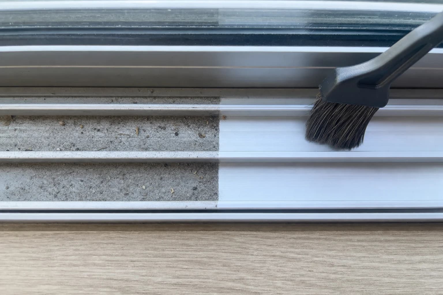 A sliding door track being cleaned with a detail brush, grit still visible in the untouched half