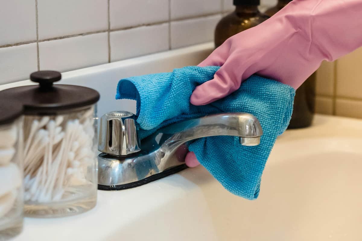 A person wearing pink rubber gloves cleaning a bathroom sink faucet with a blue microfiber cloth.